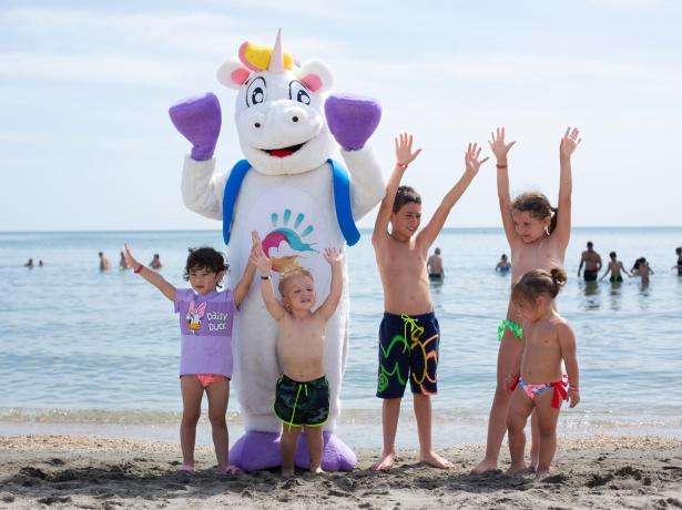 Bambini e un unicorno sulla spiaggia alzano le mani felici.