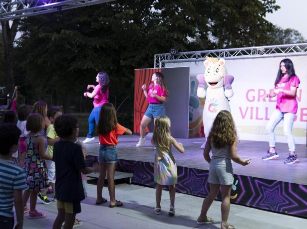 Children dance in front of a stage with entertainers and a mascot.
