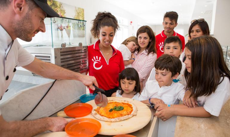 Chef taglia la pizza mentre i bambini guardano curiosi.