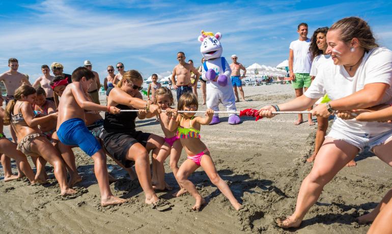 Gara di tiro alla fune sulla spiaggia con pubblico e mascotte.