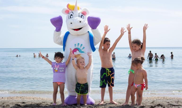 Bambini e un unicorno sulla spiaggia alzano le mani felici.