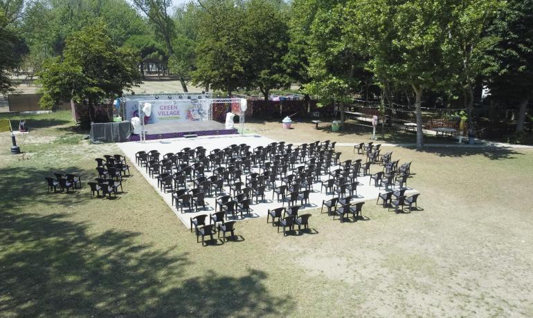 Spazio all'aperto con sedie e palco, circondato da alberi.