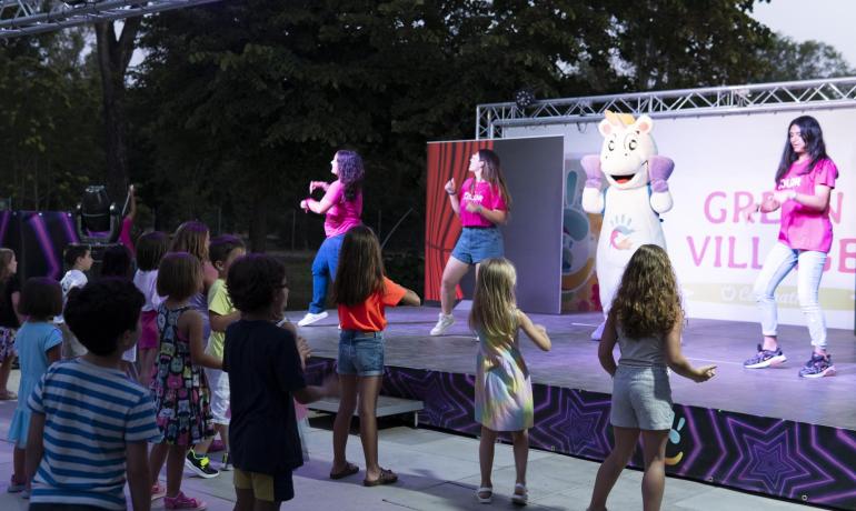 Les enfants dansent devant une scène avec des animateurs et une mascotte.