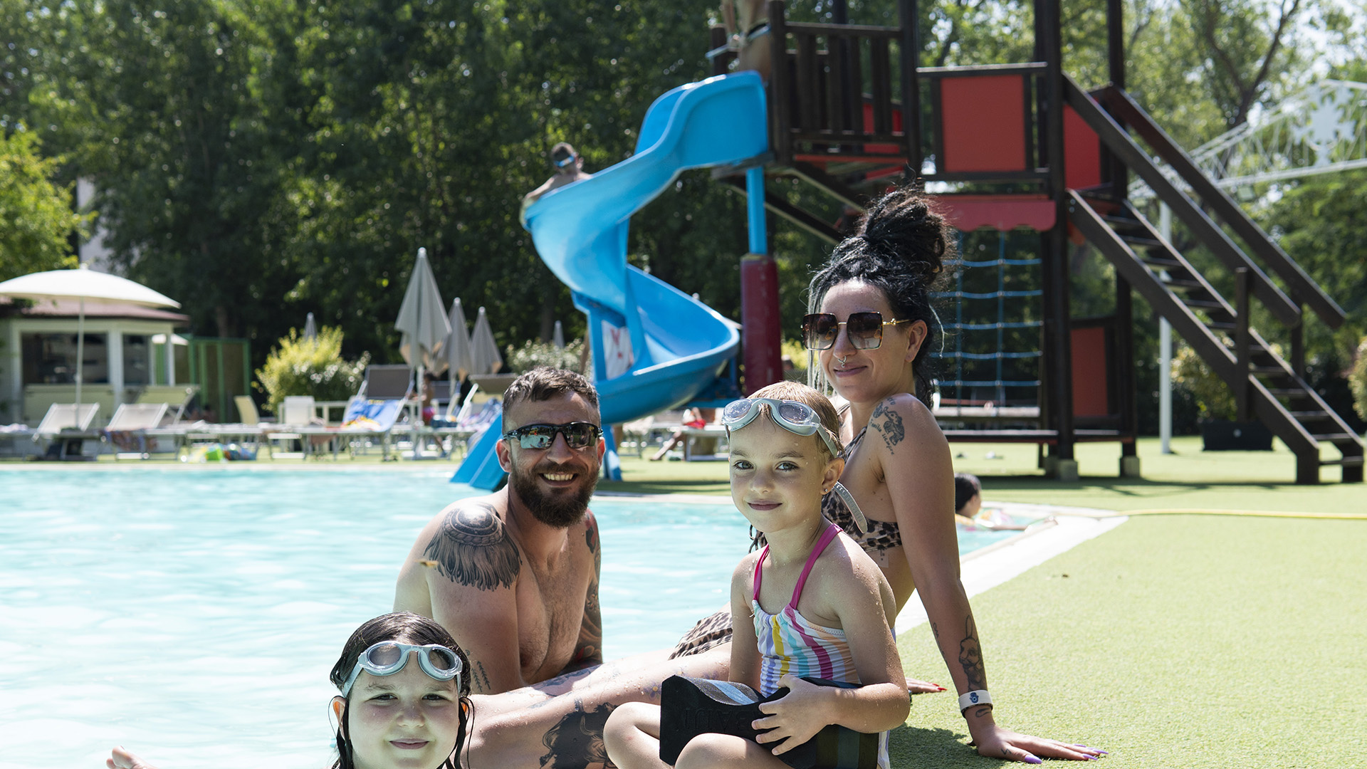 Bambino e uomo in piscina con braccioli, ombrelloni colorati sullo sfondo.