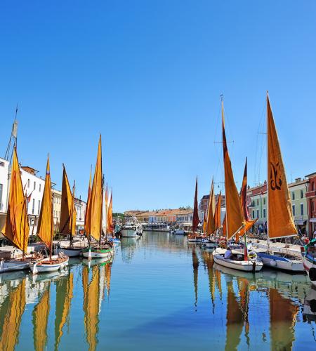 Colorful boats with orange sails reflect on the canal, clear sky, vibrant houses.