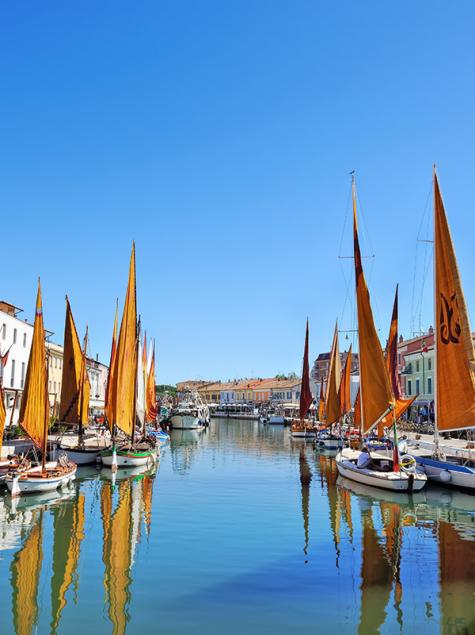 Colorful boats with orange sails reflect on the canal, clear sky, vibrant houses.